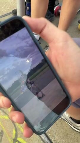 Here's a description of the video:

The video shows a person holding a phone displaying a recording of someone falling while skateboarding in a skatepark. The person on the skateboard appears to be attempting a trick on the edge of a bowl, then falls down onto the bottom of the bowl. After the clip ends, the video shows three young people watching the clip. One has curly blond hair, glasses, and is wearing a gray Nike shirt and patterned pants. He is laughing and clutching his arm. Another is sitting on a scooter wearing a pink helmet and a similar gray shirt. The third is standing, wearing a black t-shirt and light wash jeans, and appears to be holding a scooter. They seem to be at the skatepark where the fall occurred. The skatepark has graffiti on some of the ramps. The sky is cloudy in the video recording.