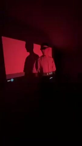In a dimly lit room, a person stands in front of a projector screen. The room is illuminated with shades of blue and purple. The projector displays a bright, colored image. The person is wearing a light-colored shirt and dark pants. The person's shadow is cast on the screen. The person puts on sunglasses.