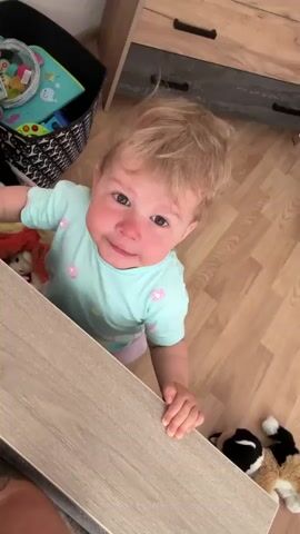 A blond-haired baby with blue eyes and rosy cheeks, wearing a light blue t-shirt with pink flower patterns and pink socks, stands at a wooden table. The baby is looking up with an upset expression and appears to be crying. The baby's hands are on the edge of the table, and the baby is leaning forward. In the background, there is a black basket filled with toys, a wooden dresser with drawers, and a wooden floor. There are also stuffed animals on the floor near the baby. The lighting suggests an indoor environment.