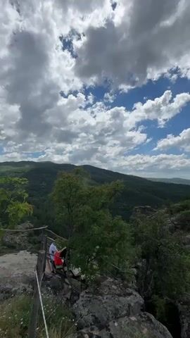 The video shows a panoramic view of a mountainous, forested landscape under a partly cloudy sky. In the foreground, there are rocks and some vegetation. On a wooden platform to the left, two people are visible, one wearing a red shirt and the other a white shirt and dark pants, both appearing to be sitting and observing the view. The camera pans across the vista, revealing rolling hills and distant mountain ranges covered in dense green forests. The overall scene depicts a natural outdoor environment, likely a scenic overlook or hiking trail.