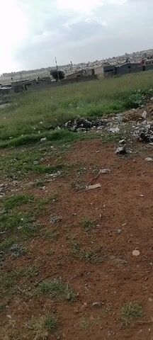 The video shows an open field with red dirt, grass, and scattered trash. In the background, there are houses and a cloudy sky. No people are visible. The camera is handheld and mostly still, with slight movements. The scene is unremarkable, depicting a neglected urban periphery.