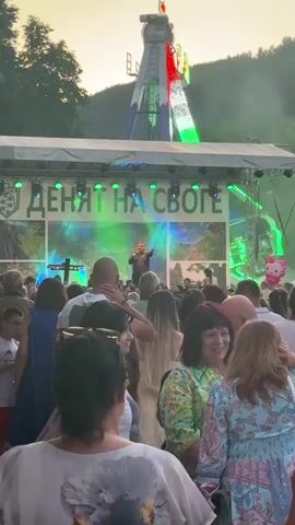 A group of people are gathered in an outdoor area, facing a stage where a performer is singing and playing an instrument. Behind the stage, a large amusement park ride with flashing green and blue lights is visible. The sky is overcast with a hint of sunset. The crowd appears to be enjoying the performance, with some individuals holding up their phones to record. A Hello Kitty balloon floats near the stage. The overall atmosphere is festive and lively.