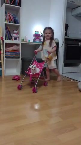 A young girl with brown hair pulled back in pigtails, wearing a pale yellow dress with light blue and pink polka dots, stands and smiles at the camera, holding the handles of a pink toy stroller. A white kitten with gray markings is nestled in the seat of the stroller. The girl is in a room with light brown hardwood floors. To the left of the girl is a white bookshelf filled with colorful books and toys. Behind her, a tall, white tower fan is visible. To the right, a kitchen area with cabinets and a stove is partially visible. The girl begins to push the stroller forward, and the kitten looks down.