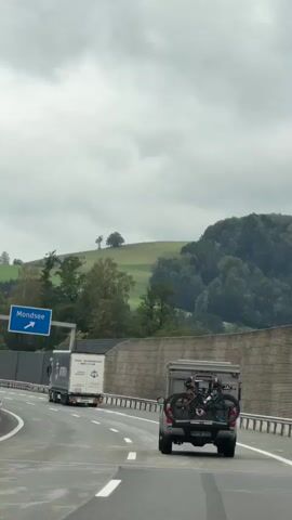 This is a vertical, first-person perspective video taken from a vehicle traveling on a highway. The camera is focused on the road ahead. In the foreground, the rear of the vehicle the camera is in is visible, including a bike rack mounted to the back with two bicycles attached. To the right of the lane, a concrete barrier lines the highway, followed by a wooded hillside. Overhead, highway signs indicate directions. One sign shows an arrow pointing up for A1, E55, E60, Salzburg, and Thalgau. Another blue sign with a white arrow pointing right indicates Mondsee. Several trucks are visible on the highway, traveling in the same direction as the camera's vehicle. The sky is overcast.