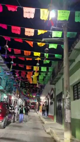 The video shows a narrow street decorated with colorful banners strung overhead. These banners are various shades of red, orange, yellow, green, and purple. Underneath the banners, there are multiple people walking along the street. Cars are parked along the side of the street. The buildings flanking the street are painted in different colors, some in muted shades of green. The video appears to be taken at night, with the street lit by streetlights.