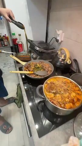 The video shows a close-up view of a kitchen stovetop where two dishes are being prepared. In the foreground, a large pot contains a bubbling, reddish-orange stew with visible chunks of food. To the left, a frying pan holds pieces of meat and green vegetables, with a spatula resting inside. A person's hands are visible using tongs to add more ingredients to the frying pan, and a pair of yellow-handled scissors are also being used. The stovetop appears to be an induction cooktop with illuminated control buttons. In the background, a black electric kettle with a yellow handle is visible, along with various bottles and containers on a shelf, including what appears to be a can of Coca-Cola. The overall environment suggests a home kitchen during meal preparation.