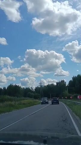 The video shows a first-person perspective of a vehicle driving on a paved road. The sky is blue with fluffy white clouds. On the right side of the road, there are trees and curved traffic signs indicating turns. Power lines run overhead. Several vehicles are visible on the road, including a dark colored minivan, a dark colored sedan, and a white semi-truck pulling a large white tanker trailer. The tanker trailer has text on its side. The vehicle from which the video is recorded has a dashboard visible at the bottom.