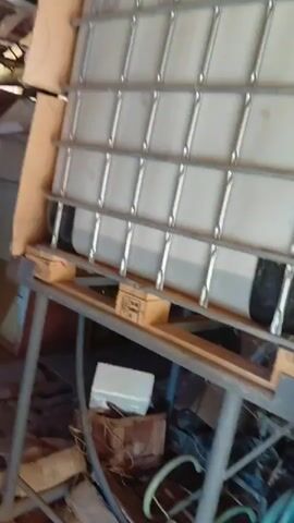 The video shows a large white plastic tank with a metal cage surrounding it. The tank is partially filled with a light-colored liquid. It is elevated on a wooden pallet, which is then supported by a metal stand. The background appears to be an indoor storage area with wooden walls and ceiling. Various objects are visible on the floor beneath the tank, including boxes, a white electrical device, and hoses. The lighting is dim.