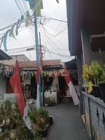 A street-level, medium shot captures an alleyway decorated for a celebration. Two Indonesian flags, one on the left foreground and another to the right, are prominently displayed, their red and white colors vibrant against the overcast sky. A cluster of colorful, narrow banners hangs across the street, adding a festive atmosphere. A utility pole with a chaotic tangle of wires rises in the center, adorned with solar panels at its apex. Several houses with tiled roofs line the alley, their facades showing signs of everyday life with plants in pots and various household items. In front of one of the houses, two individuals are seated, their interaction obscured by the distance and focus on the flags and decorations. The overall scene suggests a community preparing for or celebrating a national holiday, with a focus on patriotic symbols amidst a lived-in urban environment.