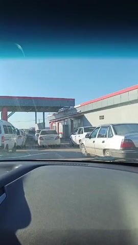 The video shows a line of cars at a gas station. The view is from inside a car, looking out through the windshield. There are several white cars and a white van in the line ahead. The gas station has a red and grey roof with grey walls. The sky is blue. The dashboard of the car in which the video is recorded is visible in the foreground. A voice in the background says that they are at a gas station and that there are 403 cars.