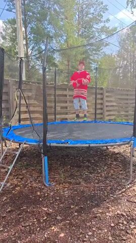 A young person wearing a red hockey jersey, a red baseball cap, and patterned shorts is jumping on a trampoline outdoors. The trampoline has a blue mat and a black safety net. The person is in the center of the trampoline, with trees and a wooden fence in the background. The ground around the trampoline is covered in brown leaves and dirt. The person jumps and moves their arms, possibly mimicking a hockey player.