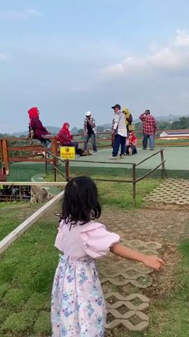 The video shows a young girl, seen from the back, walking on a stone path in a grassy area. She is wearing a light pink blouse and a colorful floral-patterned skirt, with white shoes. The path leads towards a slightly elevated platform with a wooden railing, where a group of people are standing, sitting, and conversing. Some are wearing head coverings. A sign with text is visible on the railing. In the background, there is a view of green fields and distant hills under a cloudy sky. The camera follows the girl as she walks toward the platform. A boy walks across the platform in the distance.