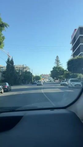 The video shows a view from inside a car looking forward through the windshield. The car is on a paved road with lane markings. The road appears to be a street in a town or city, with buildings and trees lining both sides. The sky is clear and blue, indicating a sunny day. There are other cars on the road, and some buildings under construction are visible in the distance. A tall, slender tree stands prominently on the right side of the road. Power lines are visible overhead. The video captures a brief moment of driving through this environment.