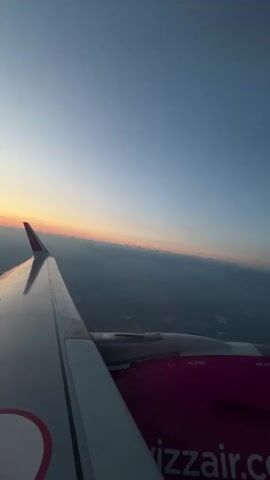 The video shows a view from an airplane window during sunset. The wing of the airplane is visible on the left side of the frame, and the fuselage with the Wizz Air logo is on the right. The sky is clear blue with a gradient of orange and pink near the horizon, indicating sunset. Below the airplane, there are clouds or mist covering the landscape, with some hints of land features. The perspective suggests the airplane is in flight.