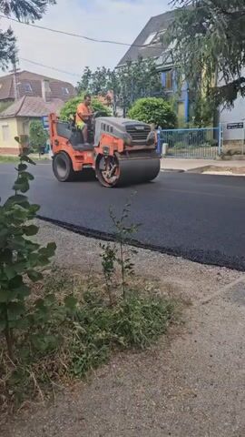 In Hévíz, Hungary, a man operates an orange and grey Hamm asphalt roller on a freshly paved road. He is wearing an orange t-shirt and green shorts. The road surface is black and appears to be newly laid asphalt. The roller is compacting the asphalt, smoothing it out. In the background, there are houses with tiled roofs and trees. A bus stop sign is visible on the left side of the road. The sky is partly cloudy, suggesting it is daytime. The scene depicts road construction work in a residential area. The text "HAMM" is visible on the side of the roller.
