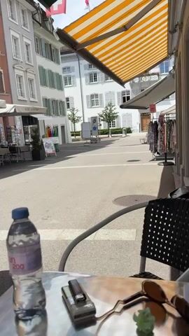 This media file captures a daytime scene at an outdoor cafe in Liestal, Switzerland. The focus is on a table set for a patron, with a bottle of Evian water, a lighter, sunglasses, and a small green plant. A black woven chair stands nearby, casting a patterned shadow on the paved ground. In the background, buildings with traditional European architecture, featuring light-colored facades and green shutters, line a street. Storefronts and awnings are visible, suggesting a commercial area. The bright sunlight indicates it is likely midday or early afternoon, and the clear sky points to fair weather. The overall mood is relaxed and leisurely, hinting at a moment of respite. Visible text includes "Evian 200" on the water bottle and "SCHÜTZENHAUS" on a building in the background. The scene conveys a sense of enjoying a calm moment outdoors in a charming European town.