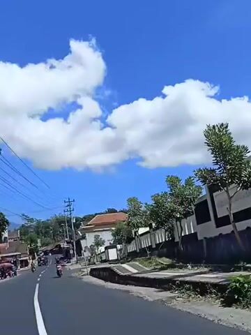 The video captures a road scene under a clear blue sky dotted with white clouds. A dark asphalt road curves to the left, with a white dashed line marking the lane. On the left side of the road, several motorcycles are visible, some with riders. Buildings line the left side of the road, appearing to be shops or houses, with utility poles and wires overhead. On the right side of the road, a white wall with black accents runs along a slight incline. Trees are planted behind the wall. The overall impression is a daytime scene in a rural or semi-urban area, possibly in a tropical region given the vegetation and the bright sunlight.
