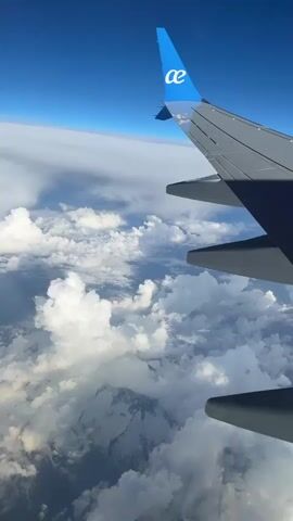 The view from an airplane window shows the wing of the plane against a backdrop of white clouds, a snow-covered mountain, and a clear blue sky. The airline logo is visible on the tail fin, and the cloud cover is thick, with a mountain peaking through.