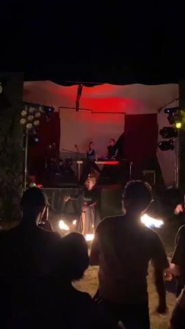 A group of silhouetted people stand in the foreground, facing a dimly lit outdoor stage at night. On the stage, two individuals are visible behind a DJ setup and musical equipment. In front of the stage, a performer in dark clothing, possibly a dancer or fire performer, is actively engaged in an activity that involves light or flames, creating bright flashes. The stage is decorated with a draped fabric backdrop and illuminated by stage lights, some of which cast red hues. The overall atmosphere suggests an outdoor performance or concert setting.