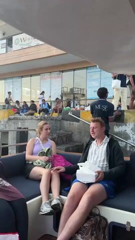 The video shows a group of young people sitting on a boat. In the foreground, a young woman with blonde hair is sitting on a dark blue seat, holding a green cloth. Next to her, a young man with light brown hair is sitting, wearing a striped shirt and holding a white container. Further down the seat, a young man with a tan baseball cap is seated, wearing a green t-shirt. A young woman with blonde hair is sitting next to him, wearing a white tank top and blue shorts. Another young woman is seated to her right, wearing a dark top. On the table in front of them, there are several bottles of water, bananas, and a box of tissues. In the background, other people are visible standing and sitting on stairs, and there is a building with signs. The boat appears to be docked or waiting.