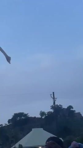 The video captures an outdoor scene in Laminga, Nigeria, likely during the late afternoon or early evening, as indicated by the soft, diffused light and the pale blue, overcast sky. The foreground is dominated by the upper portion of a motorcycle, suggesting it's either stationary or has just been moved.

In the mid-ground, several vehicles are visible, including a silver minivan and a dark-colored SUV. A person in a light-colored shirt and dark pants stands near the SUV, looking towards the right of the frame. Behind the vehicles, a hilly landscape is covered in dense green foliage and rocky outcrops. A prominent white, triangular-shaped roof structure is also visible, hinting at a building or shelter.

In the distance, power lines are strung between utility poles, crossing the frame horizontally and providing a subtle cue to the infrastructure in the area. A white piece of fabric or flag is fluttering in the upper left corner of the frame.

The overall atmosphere appears calm and relaxed, with no obvious signs of urgency or distress. The presence of people and vehicles suggests a populated area, possibly a gathering point or a roadside stop. The weather seems to be clear, albeit cloudy, with no indication of rain. The image provides a snapshot of everyday life in a rural or semi-rural Nigerian setting. No visible text is present in the video.