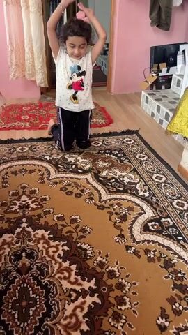 A young girl is on her knees on a rug, facing the camera. She is wearing a white t-shirt with Mickey Mouse on it and black pants. Her hands are raised above her head. She then performs a gymnastic move, flipping over and landing on her hands and feet. She is in a room with pink walls and wooden floors. There are rugs on the floor. A doorway is visible in the background.
