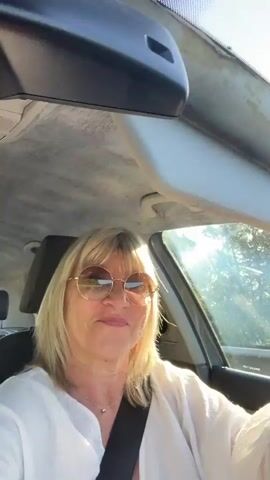 A woman with blonde hair and sunglasses is driving a car. She gives a thumbs up to the camera at 00:01. She is wearing a white top. Sunlight is coming through the window of the car.
