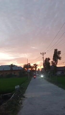 The video shows a road with a few motorcycles driving down it. The sky is filled with clouds and the sun appears to be setting, casting a pink and orange glow. There are houses and trees on either side of the road, and a field of green grass to the left. The overall scene is peaceful and serene.