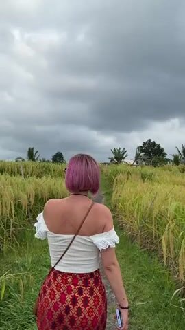 A person with short pink hair walks away from the camera down a dirt path. The person is wearing an off-the-shoulder white top and a red patterned skirt. The path is surrounded by a field of tall, yellowing grass, likely rice. The sky is overcast and cloudy.