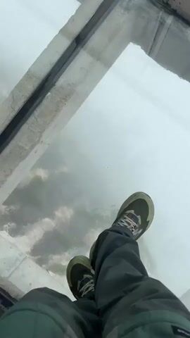 The video shows a person standing on a glass platform suspended over a foggy landscape. The person is wearing dark gray pants and light olive-colored hiking shoes with blue accents. The camera is positioned from the person's point of view, looking down at their feet on the glass surface. Below the glass, thick fog obscures most of the view, but glimpses of rocky terrain and sparse greenery can be seen through the mist. The glass platform is framed by concrete edges. The camera remains steady throughout the video, maintaining the focus on the person's feet and the foggy landscape below.