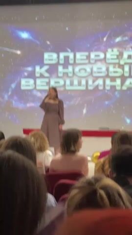 The video captures a young woman singing on a stage in front of a large screen displaying a cosmic background with the words "ВПЕРЁД К НОВЫМ ВЕРШИНАМ" (Forward to New Heights) in large, metallic letters. The woman is wearing a long-sleeved, mauve-colored dress and is holding a microphone. She appears to be performing for an audience, whose backs are visible in the foreground, seated in what appears to be an auditorium or event hall. Some of the audience members are looking towards the stage. There are some balloons, including gold ones, scattered around the stage area. The lighting suggests an indoor event, likely in the evening. The overall mood seems to be one of celebration or encouragement, as indicated by the text on the screen and the performance itself.