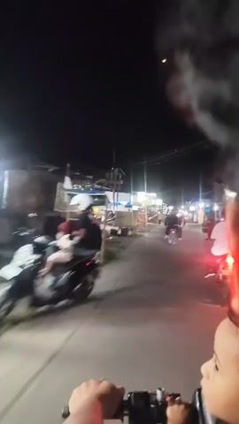 The video is filmed at night from the perspective of someone riding a motorcycle or similar vehicle. The viewpoint is slightly above and behind the handlebars, with a portion of the rider's hands and arms visible in the foreground. The road ahead is visible, with streetlights and lights from businesses illuminating the scene. Several motorcycles are also seen on the road, some with riders and some parked. Buildings and shops line the street, with signs and displays indicating commercial activity. A crescent moon is visible in the dark sky. The overall atmosphere is that of a bustling street at night.