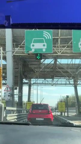 The video shows a car driving through a toll booth. The car is following a red Kia Soul. The sign above the lane shows a car with wireless symbol, and a green arrow indicating that it is OK to pass.  The traffic light is green. Signs on either side of the lane say "Backward Motion? Make sure there are no obstacles!" in Russian.  The sky is blue, and tall light poles are visible along the highway.