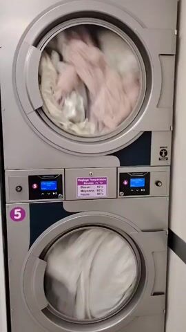 The video shows a stack of two commercial-grade dryers in a laundromat setting. The top dryer contains a load of light-colored clothing that is tumbling inside the drum. The bottom dryer also has a load of light-colored clothes. Between the two dryers is a control panel that displays the temperature settings in French: Basse (low) at 40 degrees Celsius, Moyenne (medium) at 60 degrees Celsius, and Haute (high) at 80 degrees Celsius. The dryer on the left is marked with the number 5. The environment appears to be a clean, well-lit commercial space.