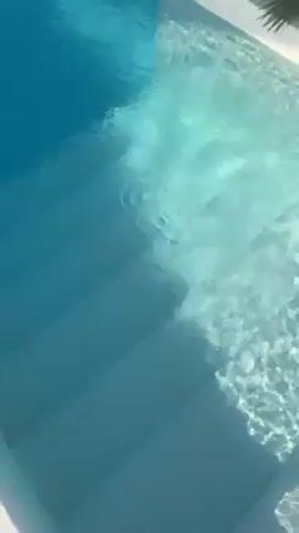 Okay, here's a detailed description of the video:

The video displays a close-up, static shot of a pool with clear, turquoise water and white steps leading into it. The water's surface shimmers with light reflections, creating a rippling effect. The steps are lined with white tiles, contrasting with the darker blue of the deeper water. A partial view of a palm tree is visible in the top right corner, providing a hint of the surrounding environment. The overall impression is one of tranquility and summer ambiance.