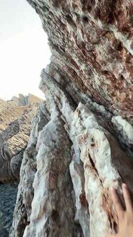 The video displays a low-angle perspective of weathered rock formations, likely along a coastline, under bright daylight. The camera pans slightly, revealing the rough, layered textures of the rocks, with streaks of reddish-brown and various shades of gray and white. In the lower portion of the frame, a person's legs and feet are visible, wearing dark Vans sneakers with white laces and soles. The person is standing on a rock overlooking a shallow body of clear, dark water that pools between the rocks, with some lighter colored sediment or algae visible beneath the surface. The water reflects the sunlight, creating a shimmering effect. The background shows more rocky terrain and a bright, overcast sky. The overall impression is one of a natural, rugged coastal environment.