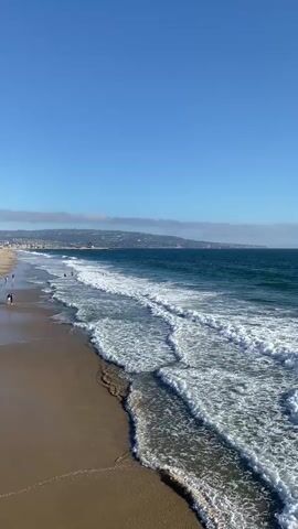 A high-angle, full shot captures a beach scene under a clear blue sky with a few wispy clouds on the horizon. The foreground shows a sandy beach with gentle waves washing ashore, creating white foam. In the distance, the ocean meets a coastline with a line of buildings and hills. A few people are visible on the beach and in the water, some appearing to be swimming or wading. The overall atmosphere is serene and coastal.