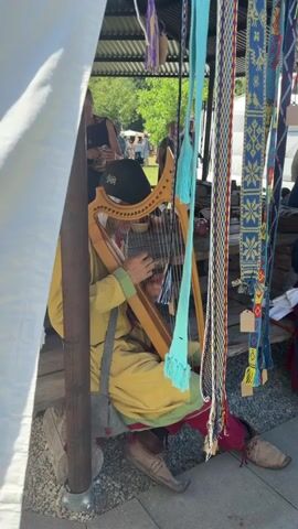 A person wearing a yellow tunic and a black baseball cap is playing a harp. They are sitting in what appears to be a market or fair, under a white tent or canopy. Behind them, several colorful woven belts or sashes are hanging. In the background, other people are visible, suggesting a public gathering. The harp is made of wood and has a number of strings. The person is using their hands to pluck the strings. The overall scene suggests a historical reenactment or a folk festival.