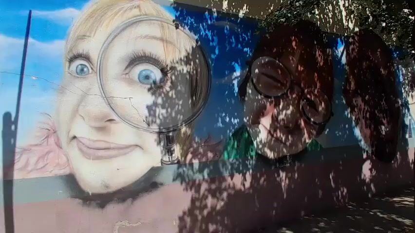 This media file is a video depicting a colorful mural painted on a wall alongside a sidewalk. The mural features large, stylized portraits of people, some of whom are looking through a magnifying glass. One portrait on the left shows a person with large, wide blue eyes and a smile, peering through a magnifying glass. The mural extends down the wall, showing more faces, many with glasses, against a blue and cloudy sky background. The artwork is vibrant and appears to be in good condition.

The scene is set outdoors, with trees casting dappled shadows across the mural and the sidewalk. The sun is shining, suggesting it is daytime, likely morning or afternoon, given the strong shadows. The sidewalk is paved with bricks, and a street with parked cars runs parallel to the wall. The architecture visible in the background, with balconies and tiled roofs, indicates an urban or town setting. The presence of parked cars and a sidewalk implies a public street.

The overall mood conveyed by the mural is one of curiosity and perhaps a touch of whimsy, particularly with the magnifying glass motif. The video pans along the wall, allowing viewers to take in the details of the artwork and the surrounding environment. The trees suggest a pleasant, possibly Mediterranean climate, consistent with the provided location context of Salt, Spain. No visible text is present in the media file.