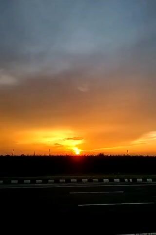 The video captures a sunset scene. The sky transitions from a deep blue and grey at the top to vibrant orange and yellow hues near the horizon. A dark silhouette of trees and vegetation forms a line across the middle ground. In the foreground, a road is visible with distinct white lines and a reflective barrier on the side. The sun is low on the horizon, partially obscured by clouds and trees, casting a warm glow. The video pans slightly to the right, revealing more of the road and the expansive sky. There are no people or animals visible in the frame. The overall environment appears to be an open, possibly rural or semi-rural area with a road infrastructure.