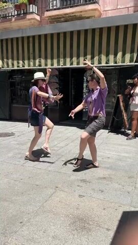 Two young women are dancing in a street in Cádiz, Spain. The woman on the left wears a white hat, a purple and pink patterned scarf over a purple top, and denim shorts. She has on a pair of white sandals. The woman on the right wears sunglasses, a purple short-sleeved shirt, and gray cargo shorts. She has on a pair of black sandals. The two women are in a side-by-side dance pose, facing each other with their arms raised and bent. Their bodies are angled, with their hips and legs extended to the side.

The dance appears to be a spontaneous and joyful expression, possibly a form of street performance or a playful interaction between friends. The women's facial expressions are not clearly visible due to distance and sunglasses, but their posture suggests energy and engagement.

The setting is a sun-drenched street paved with cobblestones. Behind the dancers is a building with a striped awning over shop windows. The building's facade is painted a pale pink. In the background, to the right, another woman is visible, holding a phone and seemingly filming the dancers. A street sign, or information board, is leaning against the building. The overall atmosphere is bright and lively, suggesting a warm, sunny day, typical of southern Spain. The time of day is likely midday or early afternoon, given the strong sunlight and shadows cast on the ground. No text is clearly visible in the media file.