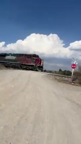 The camera captures a train crossing a rural road. The train engine is red, white, and green, with "Ferromex" written on the side. The train is traveling from right to left across the screen, moving past a railroad crossing sign that reads "Alto." The surrounding area is dry and dusty, with sparse vegetation. The sky is blue with scattered white clouds. The camera appears to be stationary, positioned on the side of the road as the train passes.