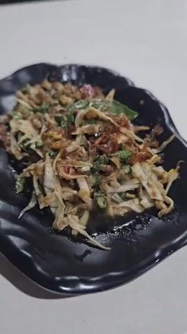 A close-up shot shows a black plate with a dish of shredded vegetables and toppings. The dish appears to be a salad or a slaw, with finely chopped cabbage, green herbs, and a reddish-brown sauce or topping. The plate is wavy-edged and rests on a white surface. The lighting is dim, suggesting an indoor setting, possibly a restaurant. No people or specific actions are visible.