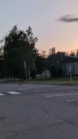 The video opens with a view of a road and a parking lot. In the background, there is a fence, a building, and trees. The sky is a soft pink and orange, suggesting either sunrise or sunset. A street lamp is visible. There is a sign with a wheelchair symbol. A small animal, possibly a deer or a large rabbit, is seen crossing the road in the middle distance. The camera then shifts, showing the interior of a car, with the side mirror reflecting the scene outside. The road and the same building and fence are visible in the reflection. The video continues to show the exterior scene.