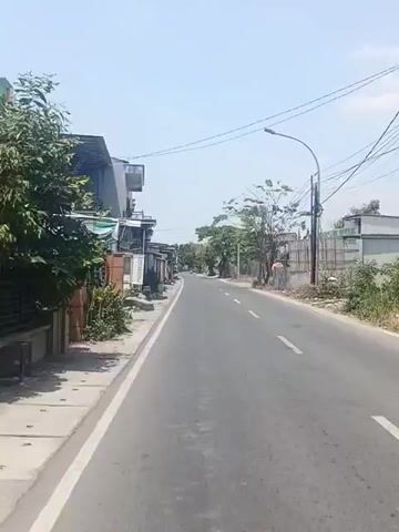 The video shows a view from a moving vehicle on a paved road under a clear sky. The road has a white line painted on the right side. On the left side of the road, there are buildings with various architectural styles and some greenery. The buildings appear to be residential or commercial structures, some with awnings and balconies. Further down the road, more buildings and trees are visible, and there is a utility pole with wires. On the right side of the road, there are more trees, a fence, and some buildings in the distance. The overall scene depicts a street in what appears to be a suburban or rural area with a moderate amount of development.