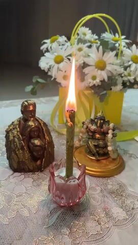 A burning green candle stands in a pink glass holder on a white lace tablecloth. To the left of the candle is a golden statue of a family embracing. To the right of the candle is a small golden statue of Ganesha, adorned with beads. In the background, a yellow basket overflows with white daisies and green leaves, tied with yellow ribbon.
