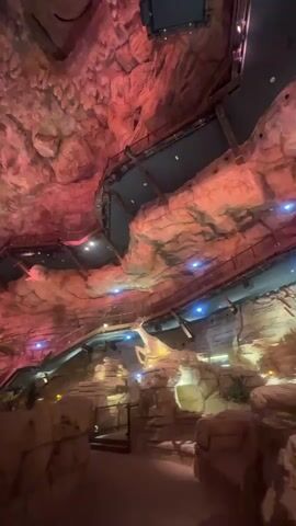 The video captures an expansive indoor cavern-like environment with intricate, artificially sculpted rock formations on the ceiling and walls. The space is dramatically lit with a shifting array of red, pink, blue, and white lights, creating a vibrant atmosphere. Multiple elevated walkways and bridges crisscross the cavern at different levels, suggesting a multi-story attraction or pathway for visitors. Various architectural elements resembling rock structures are integrated into the design. A person's head is briefly visible in the bottom right corner, looking up at the illuminated ceiling. The camera moves, panning upwards and across the scene, showcasing the scale and detail of the themed environment, which is part of Qiddiya City in Saudi Arabia.