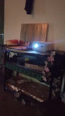 The video shows a projection of a children's cartoon on a wall. The cartoon features animated characters, including a yellow bunny, a green turtle, and a baby. The scene in the cartoon is a bright, colorful landscape with a rainbow and a smiling sun. The projection is being cast from a white projector placed on a shelf. The shelf is decorated with flowers and has various items on it, including what appears to be a canvas painting. The surrounding environment appears to be a dimly lit room.