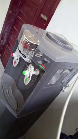 A medium shot shows a gray Arisa brand water dispenser standing against a white wall next to a dark red door. The dispenser has a water bottle receptable on top and two spigots, one with a red dot and the other with a green dot. The dispenser has a power indicator light, a standby switch, and a heating indicator light. The unit appears slightly dirty. A white power cord extends from the back of the dispenser. The camera is positioned slightly above and to the side of the dispenser. The floor is covered in white tiles. The background consists of the dark red door and white wall.