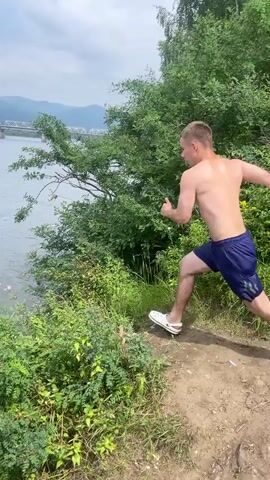 A young man is captured mid-air as he jumps from a wooded cliff into a wide river. He is wearing blue shorts and is seen in a dynamic pose with his arms outstretched. The water below him is calm with some ripples. In the background, a distant bridge spans the river, and a small pink raft is visible on the water. The environment appears to be a natural landscape with trees and hills. The video progresses as he enters the water, creating a large splash, and disappears beneath the surface amidst the churning water.
